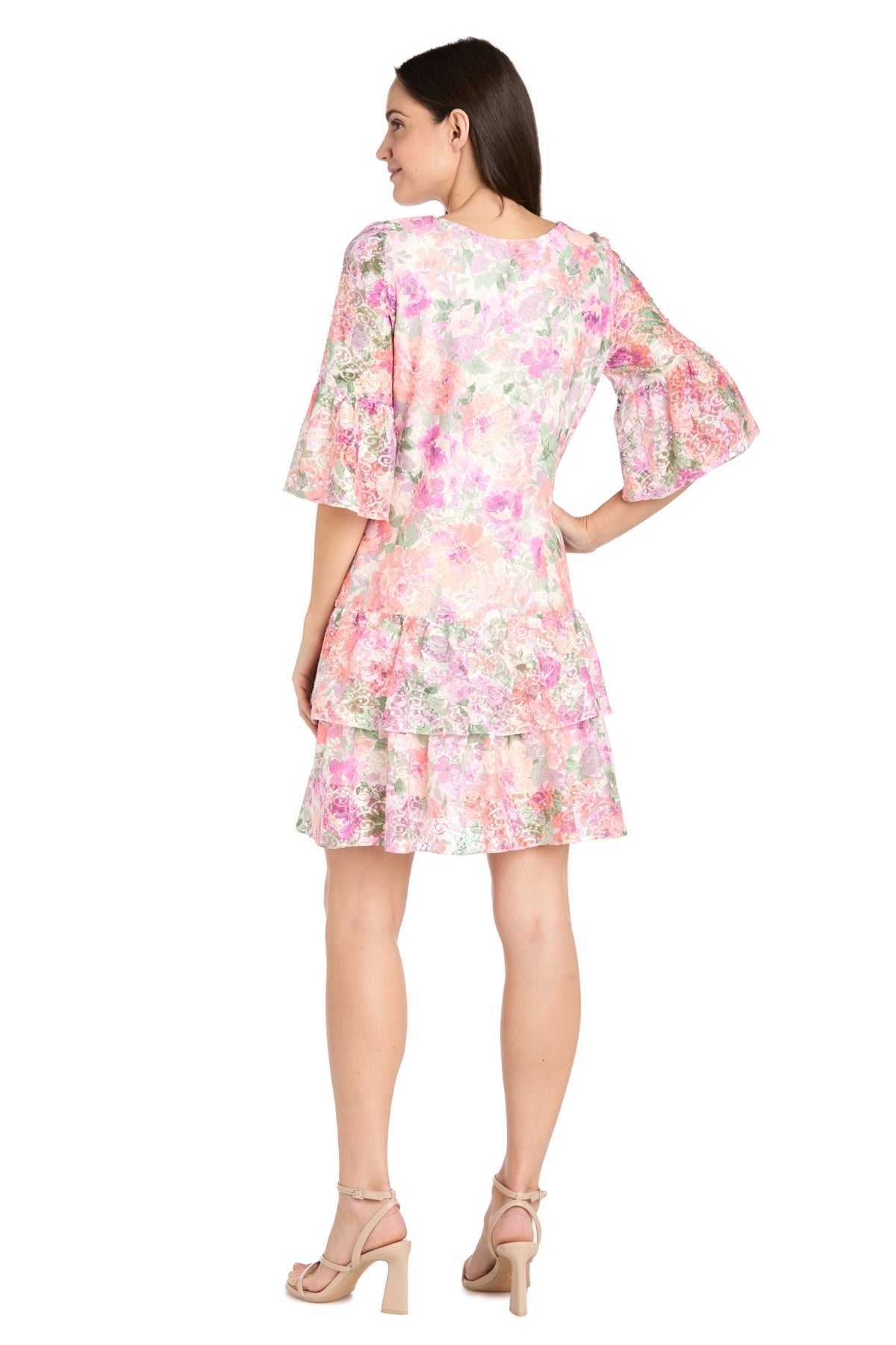 A woman faces away, wearing the Short Ivory Floral Party Dress by R&M Richards (style 4107) featuring ruffled sleeves and a layered skirt, paired with nude high-heeled sandals.